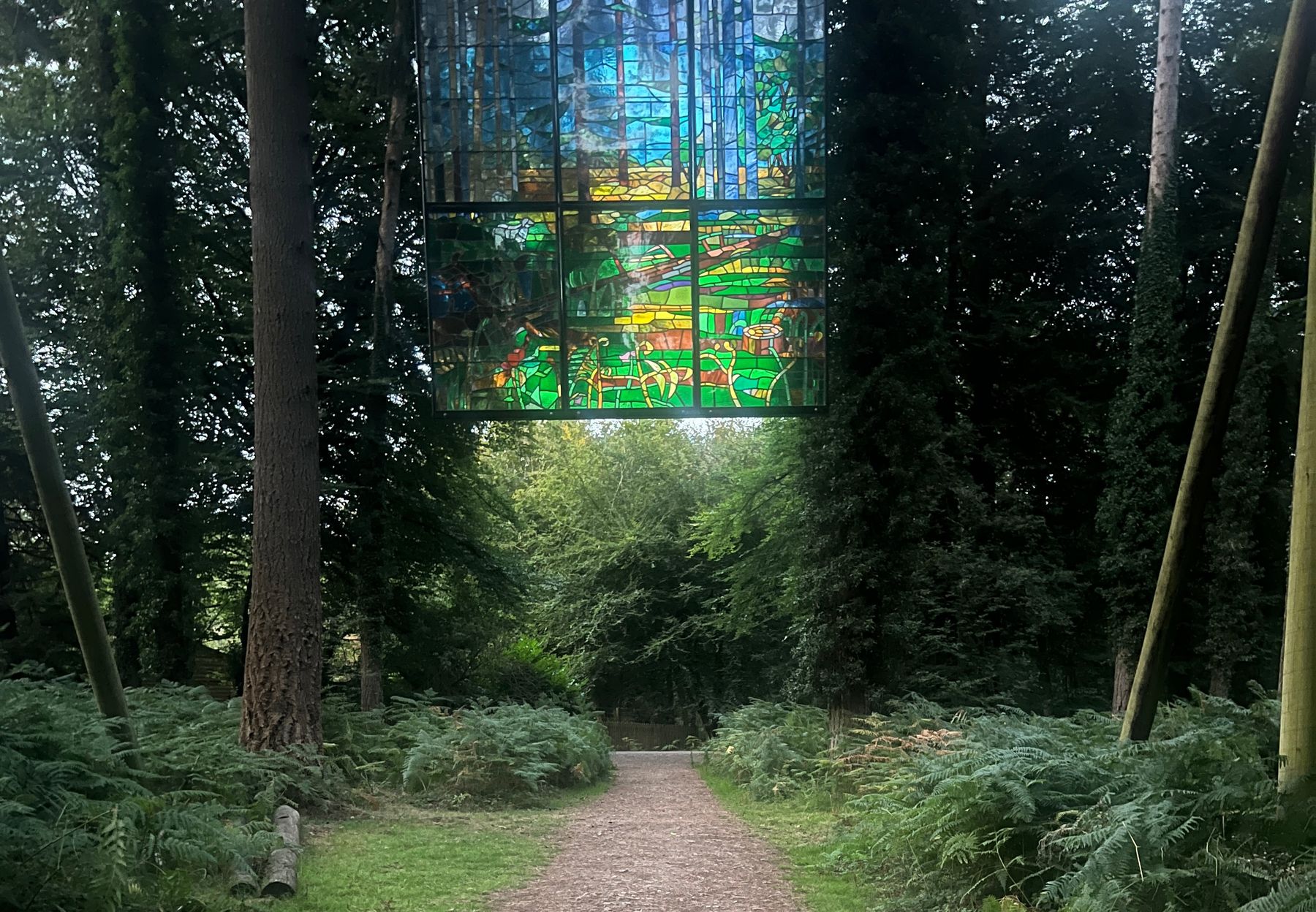A gravel path through bracken and tall conifers, with a large stained glass window hanging above it